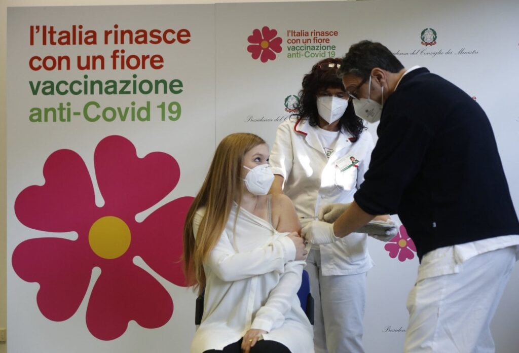 In primo piano una persona che sta per essere vaccinata, sullo sfondo pannello con la scritta "L'Italia rinasce con un fiore Vaccinazione anti-Covid 19"