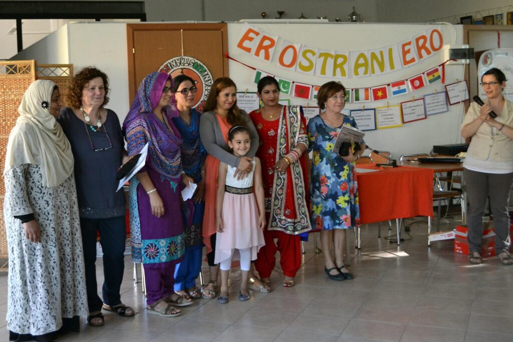 Foto di donne con il burka e volontarie. Sullo sfondo una lavagna con il volantino con la scritta "EroStraniero".