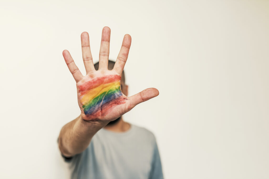 Sullo sfondo sfocato il profilo di un uomo che tende il braccio e con il palmo della mano aperta, che copre il viso, con disegnati i colori dell'arcobaleno