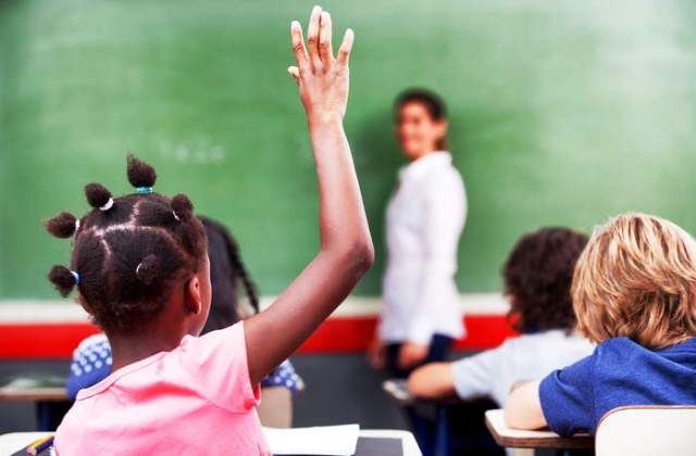 immagine di un'aula scolastica con il primo piano di spalle una bambina di colore con la mano alzata e sullo sfondo sfocato un'insegnante davanti a una lavagna. 